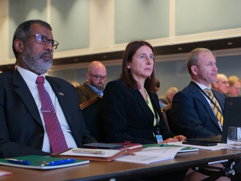 Two men and a woman, professionally dressed, listening to a lecture.