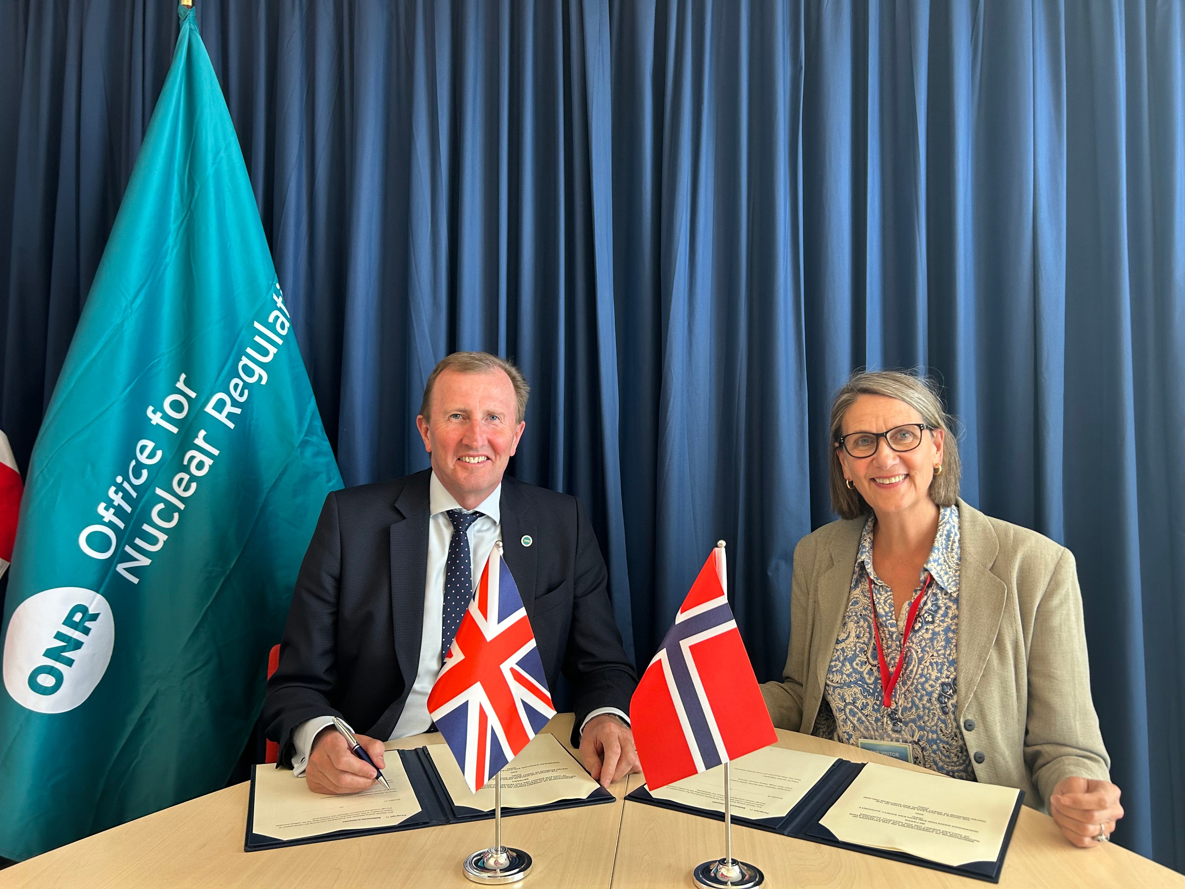 Man and woman behind a desk, with one copy each of the agreement, ready to sign. Both are smiling to the camera. 