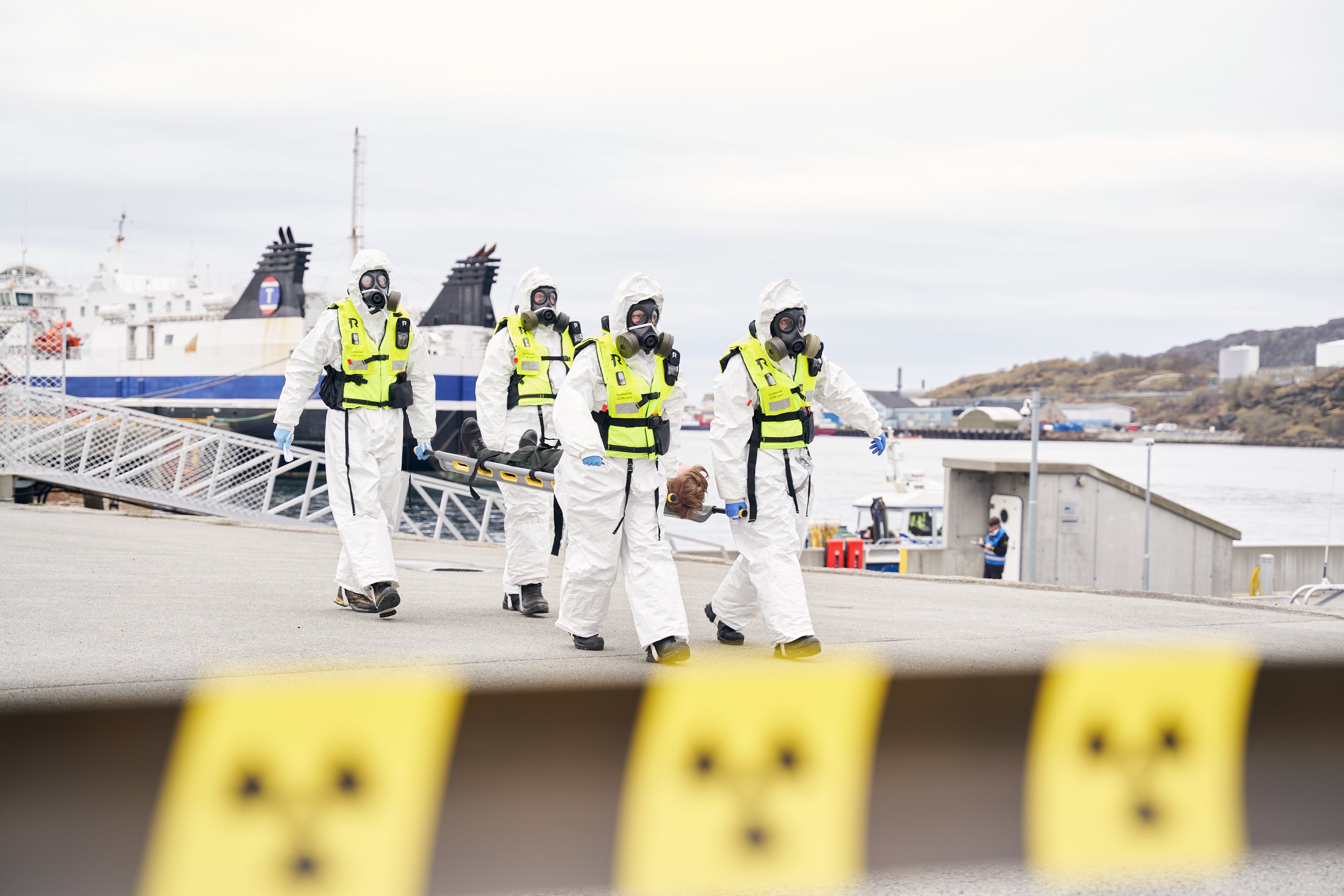 Fire personar med gassmasker og heildekkjande, kvite drakter ber ein person på båre mellom seg.