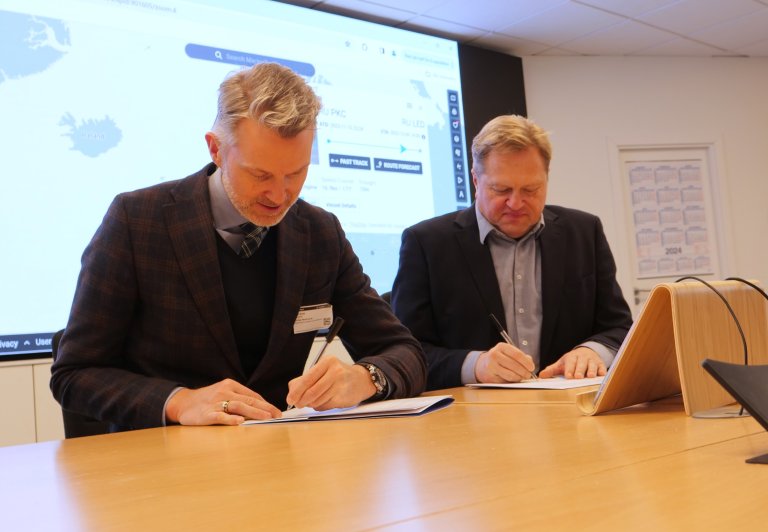 To menn i dress ved eit møtebord signerer på kvart sitt papir. I bakgrunnen ein stor skjerm med eit kart.